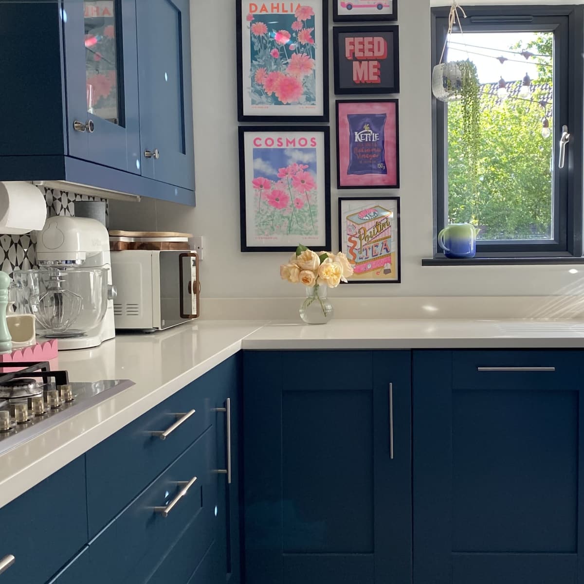 A modern kitchen featuring deep blue cabinets, a white countertop, a window with natural light, and several framed posters on the wall.
