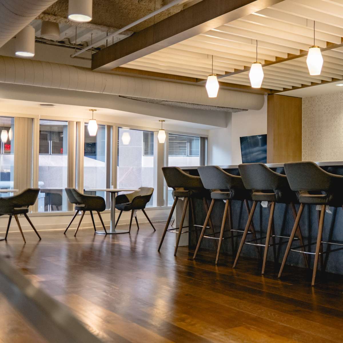A modern interior space featuring high bar stools at a counter, with pendant lights hanging above and large windows providing natural light.