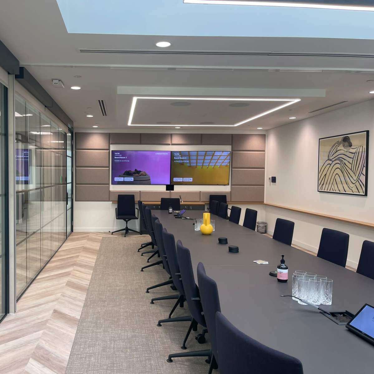A modern conference room featuring a long table, ergonomic chairs, two large screens on the wall, and glass partitioning that leads to an adjacent room.