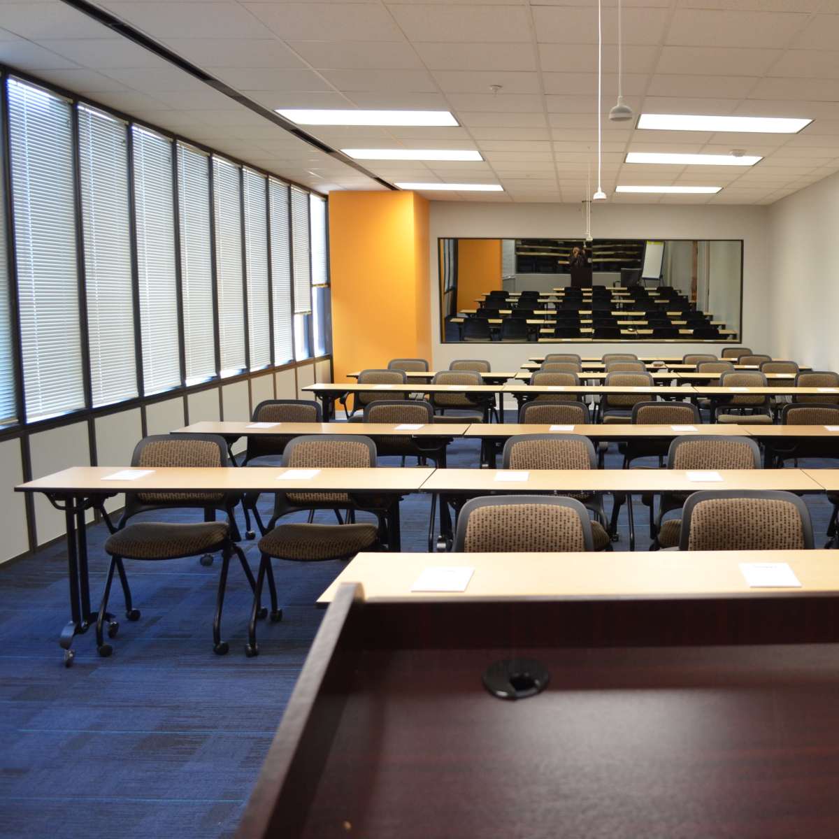 A classroom with arranged tables and chairs, featuring an empty podium in the foreground and large windows on one side.