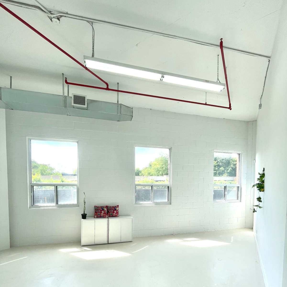 A bright, empty room with white walls, two windows, a ceiling with exposed piping, and a small table against one wall.