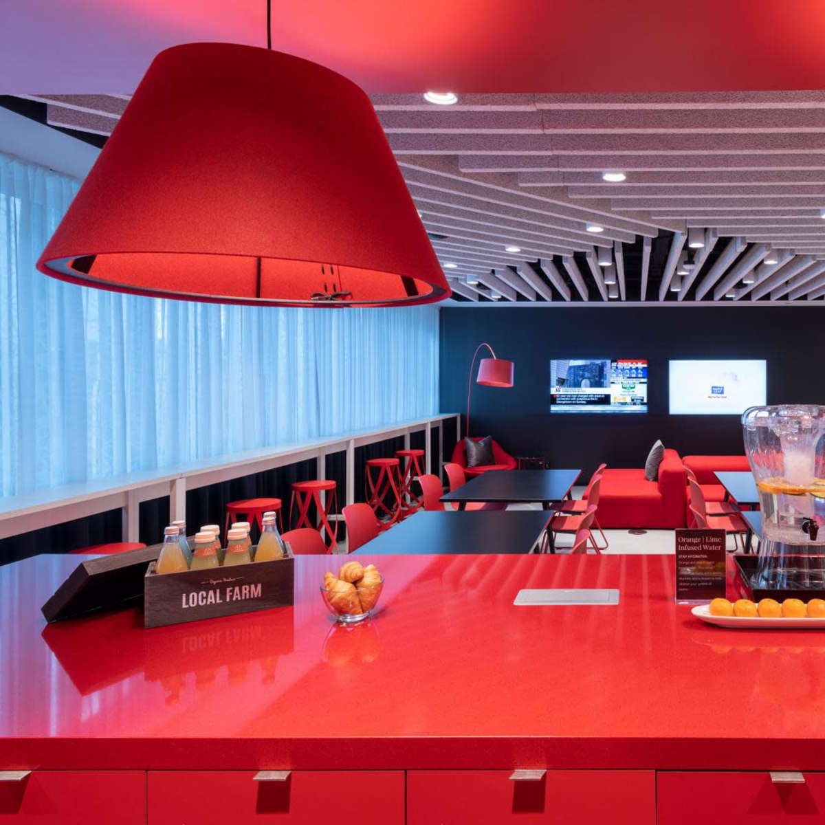 A modern lounge area is showcased with red furniture, large red lamps, and a bar counter displaying drinks and fruit.