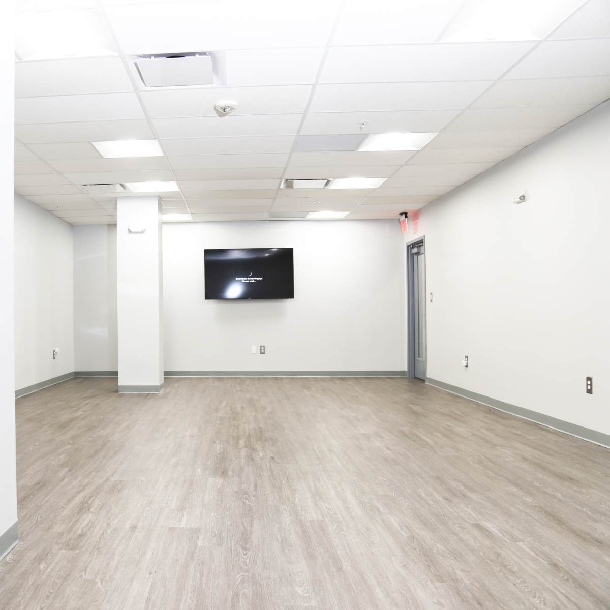 A spacious, empty office interior with light gray walls and a wooden floor, featuring a wall-mounted television.