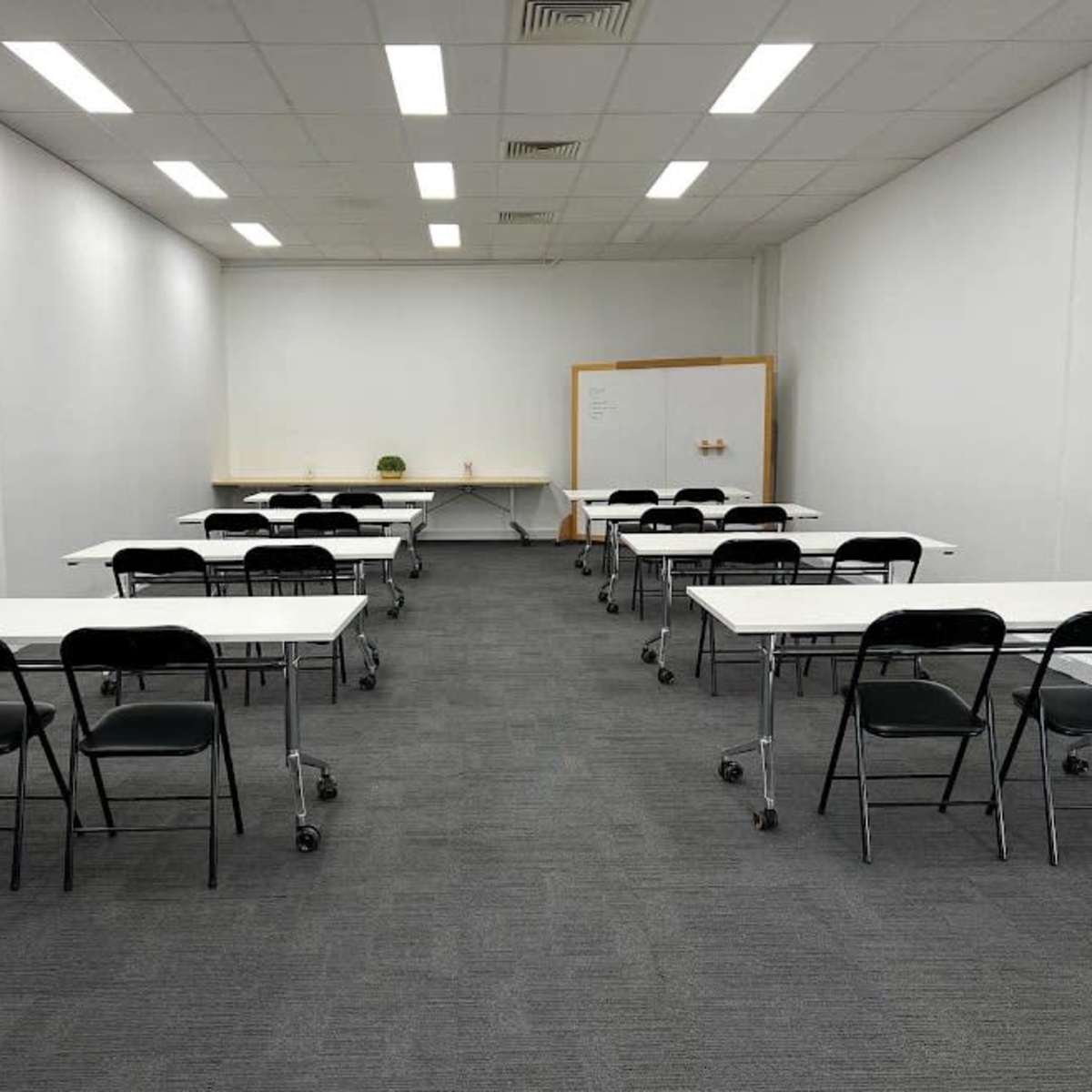 A brightly lit classroom with several rows of movable tables and chairs arranged in a neat layout against white walls.