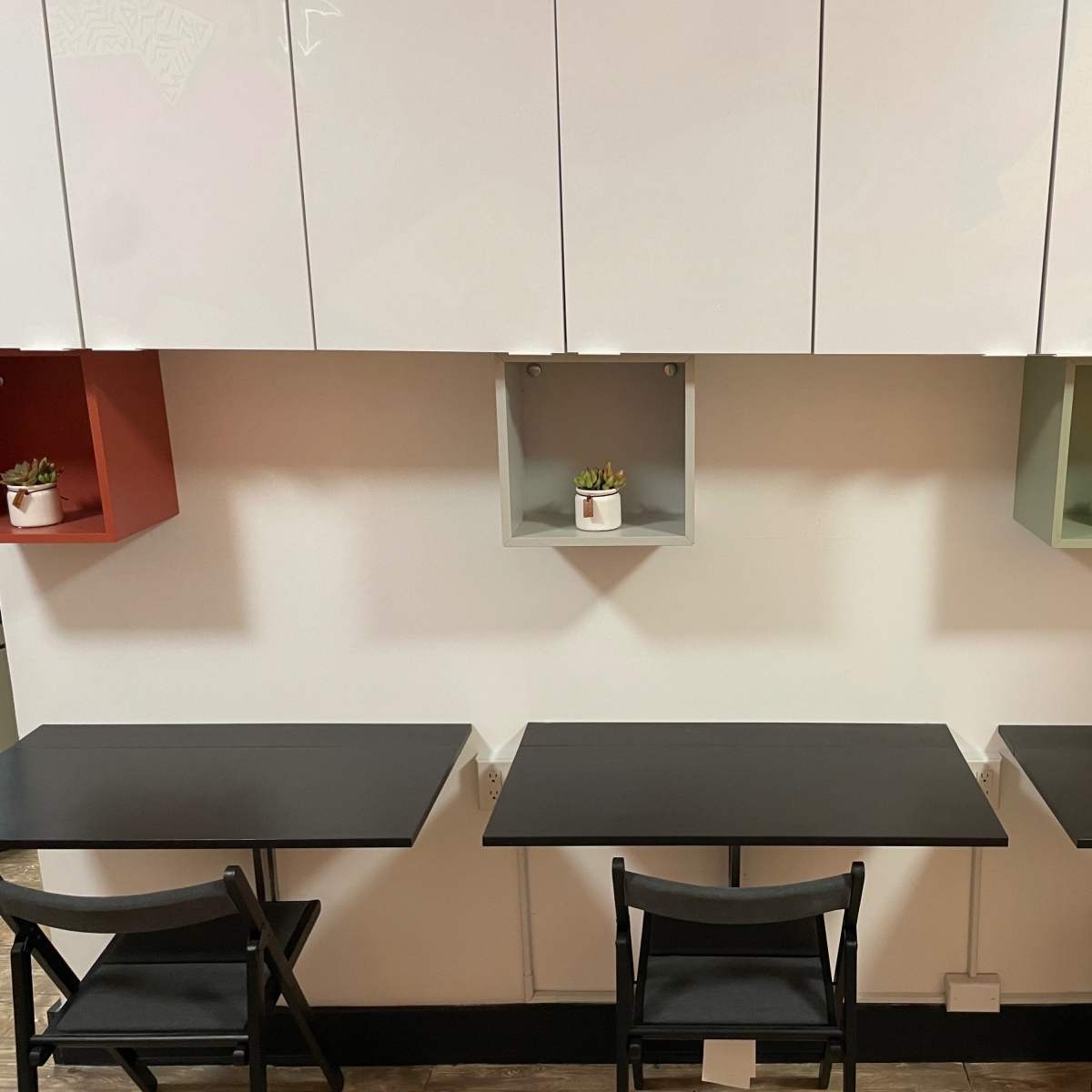 A minimalist café interior with three black tables and chairs placed against a wall featuring mounted shelves with small potted plants.