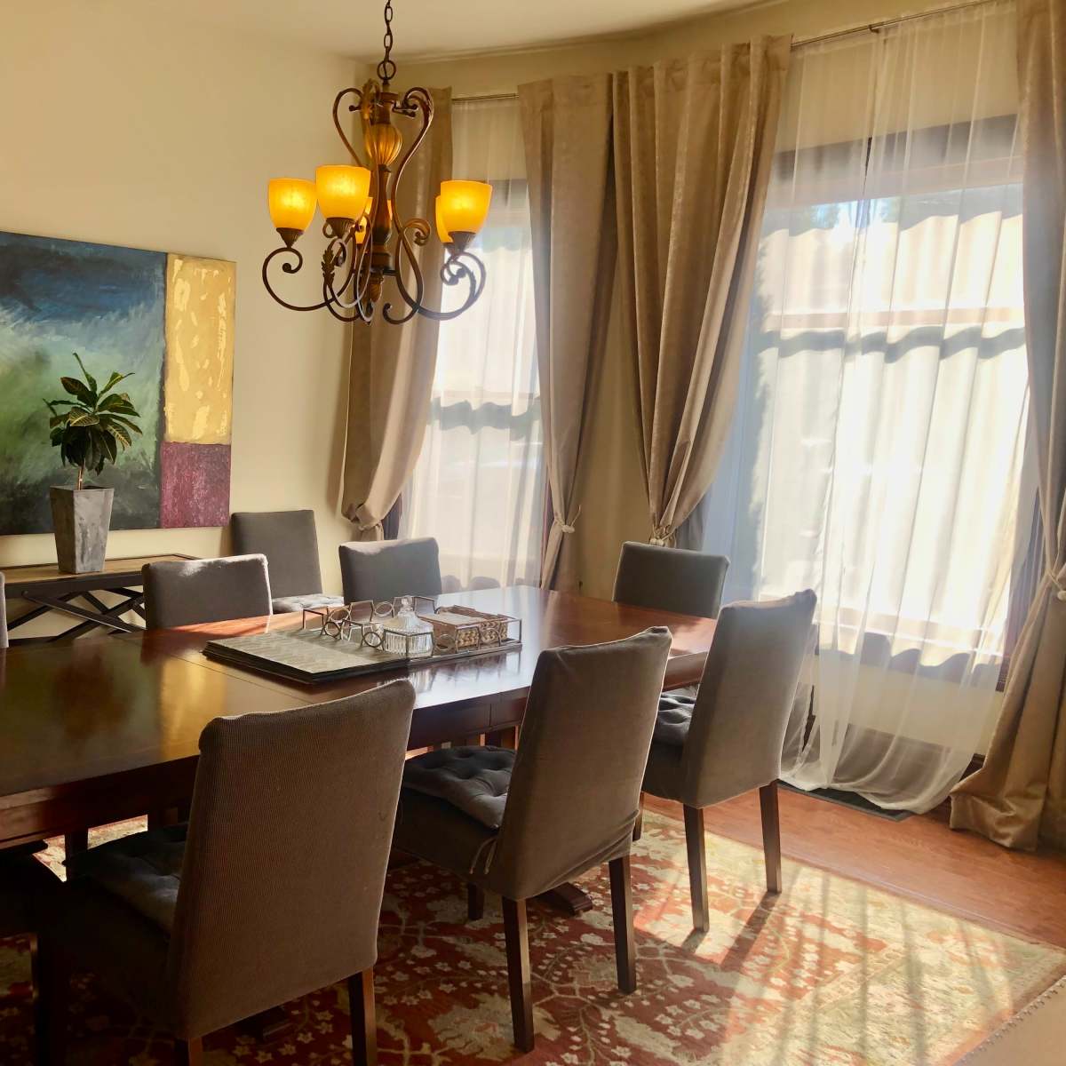 A dining room features a large wooden table surrounded by gray upholstered chairs, with a chandelier above and a colorful abstract painting on the wall.