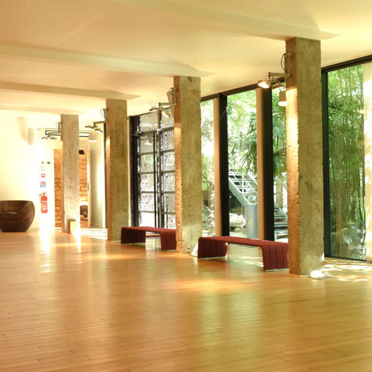 A spacious room with wooden flooring, large glass windows, and concrete pillars, featuring minimalist decor and greenery visible outside.