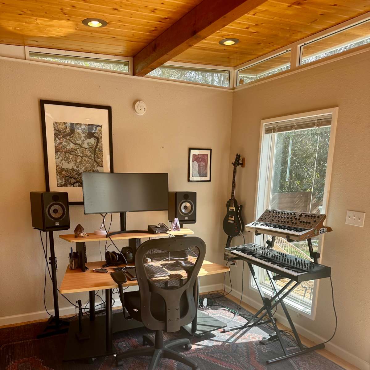 A modern music production setup featuring a desk with a computer monitor, speakers, a guitar, and a synthesizer in a well-lit room with large windows.