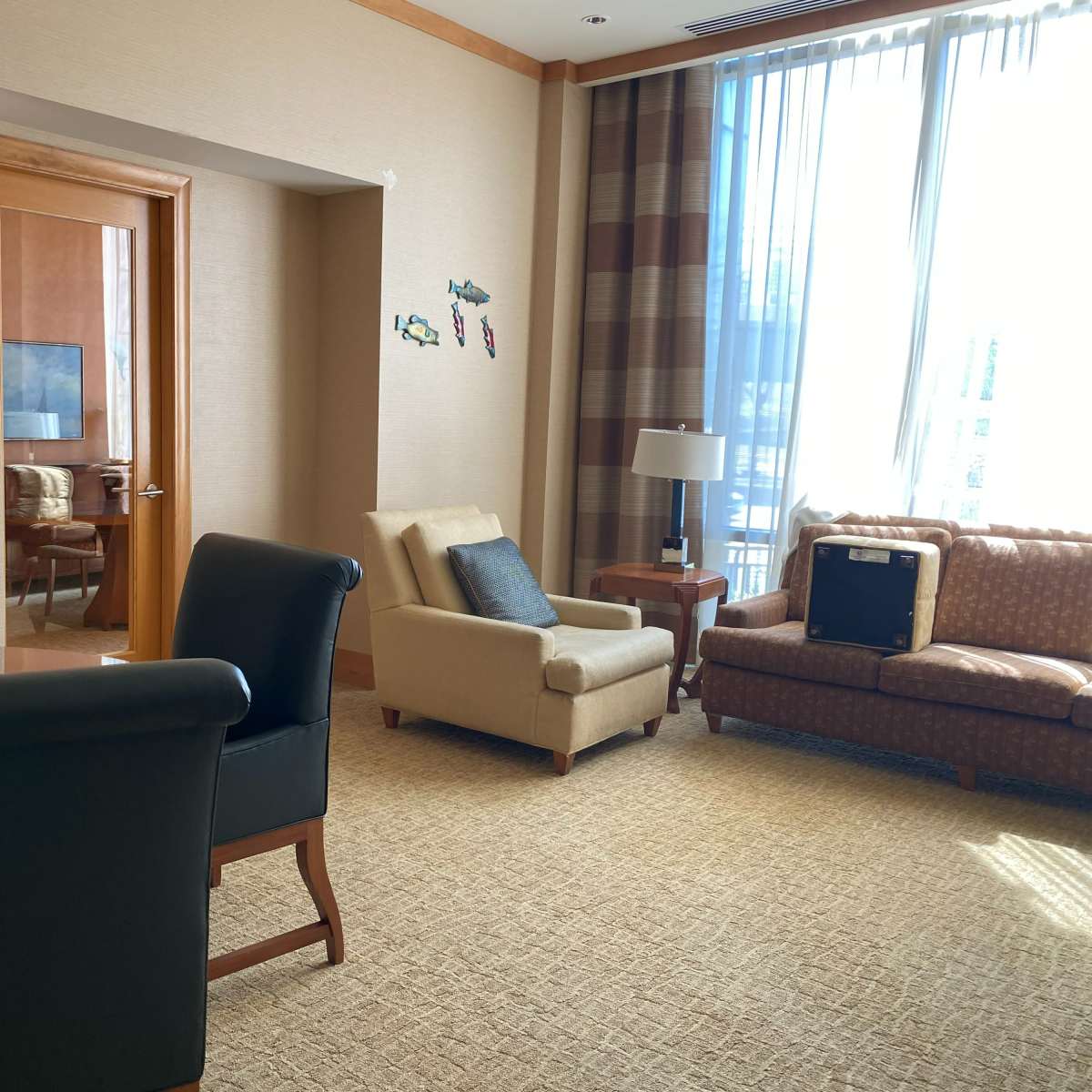 A hotel room seating area with a beige sofa, a single armchair, a round table, and large windows allowing natural light to enter.