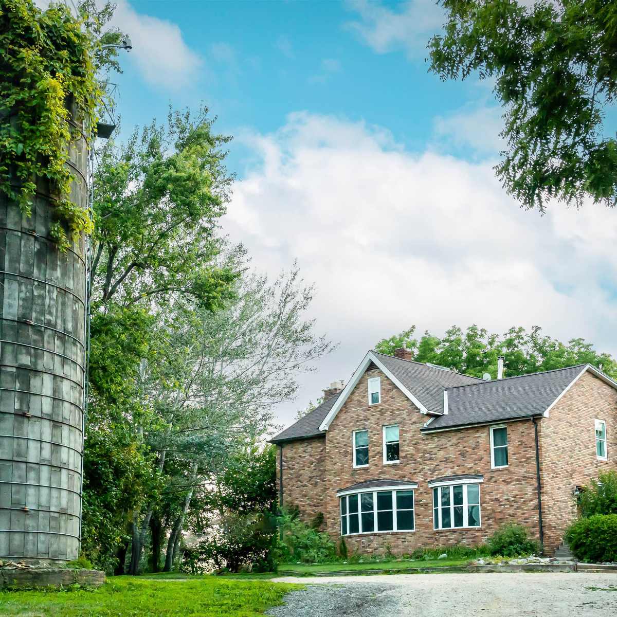 A tall, cylindrical silo covered in greenery stands next to a large, brick house with multiple windows.