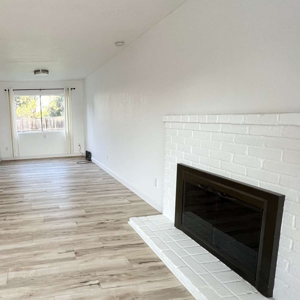 A spacious living room with light wood flooring, white walls, and a brick fireplace.