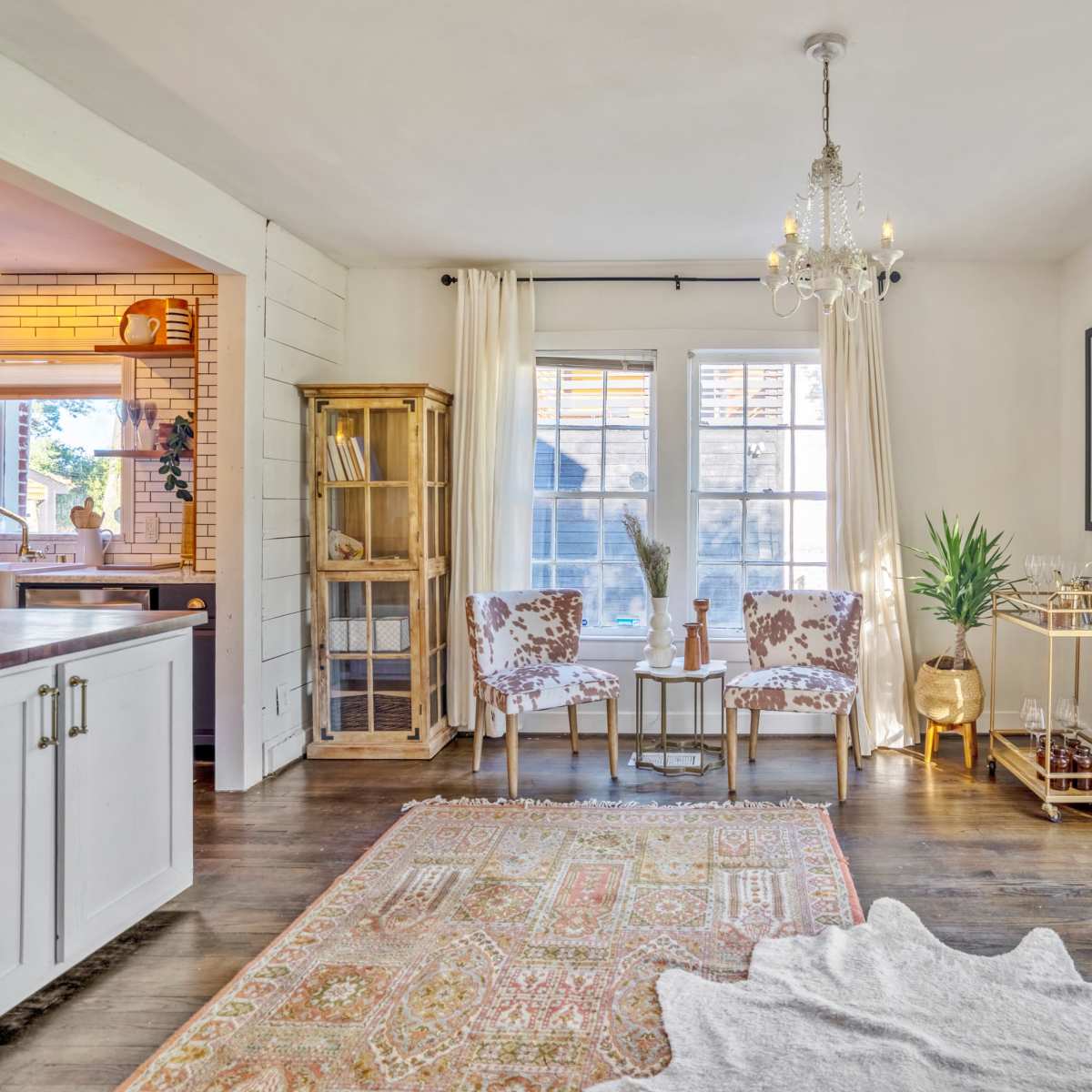 A bright living room with two chairs, a small table, a bookshelf, and decorative plants, framed by large windows and light curtains.