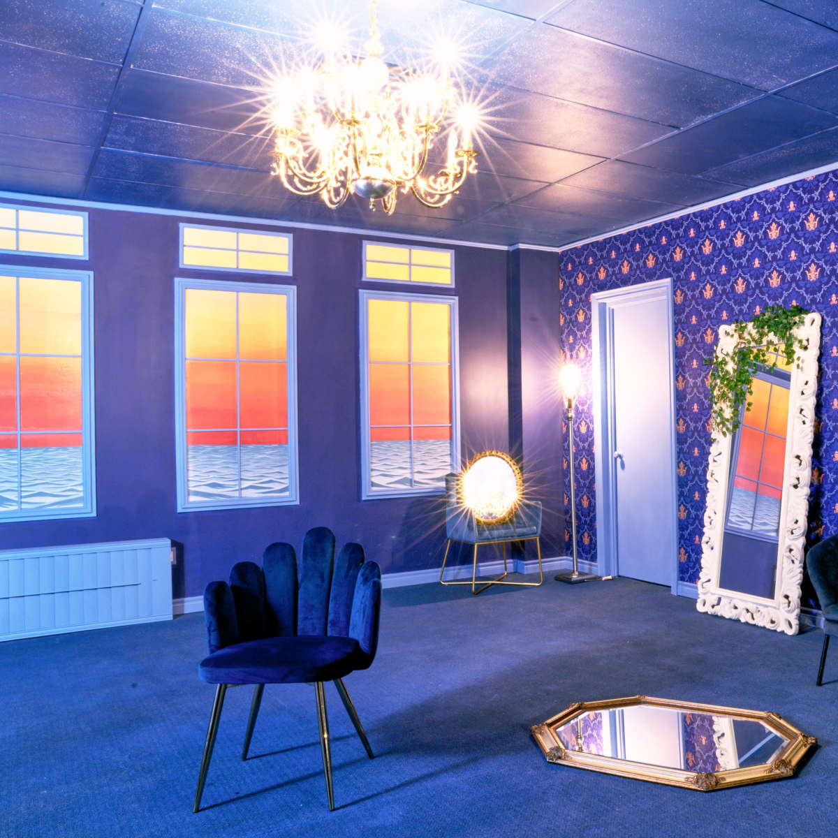 A stylish room with a patterned purple wall, a velvet sofa, a decorative mirror, a chandelier, and large windows overlooking a colorful sunset.