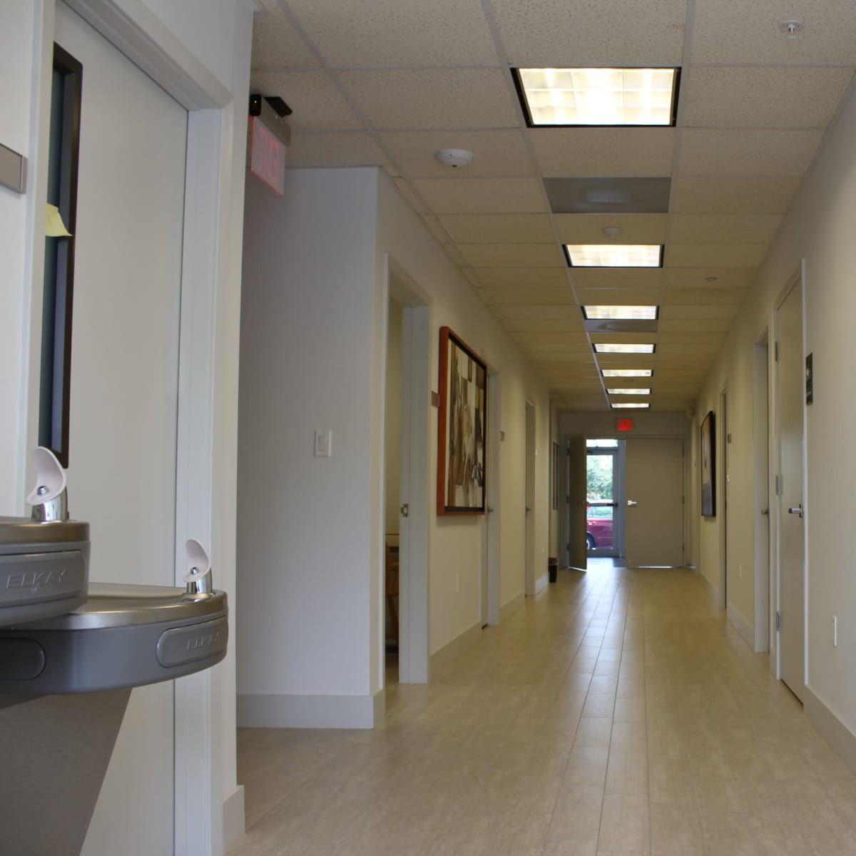 A long, brightly lit hallway featuring doors on either side and a water fountain against the left wall.