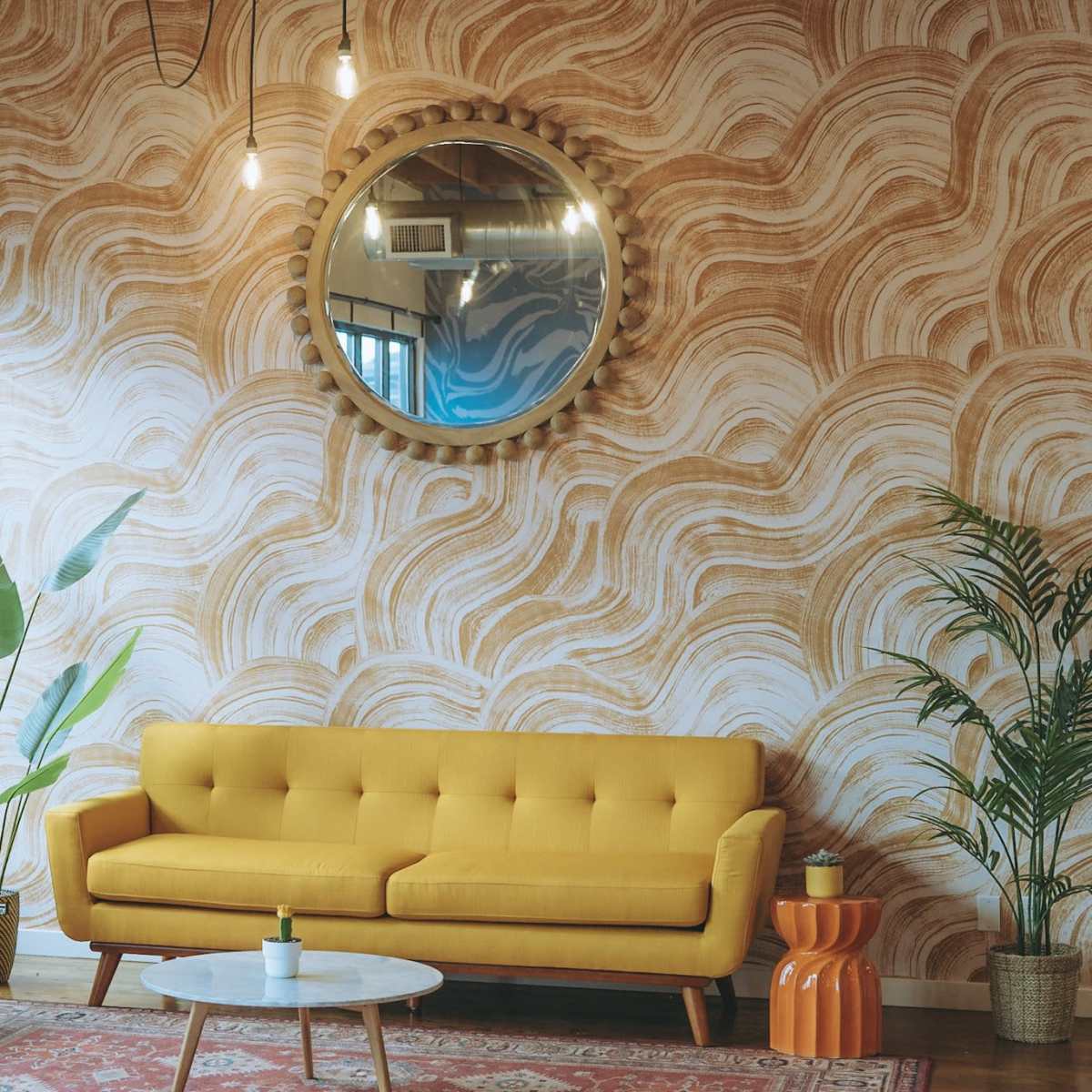 A bright yellow couch and an orange armchair are arranged in a room with a wavy patterned wall and large windows, complemented by plants and a round mirror.