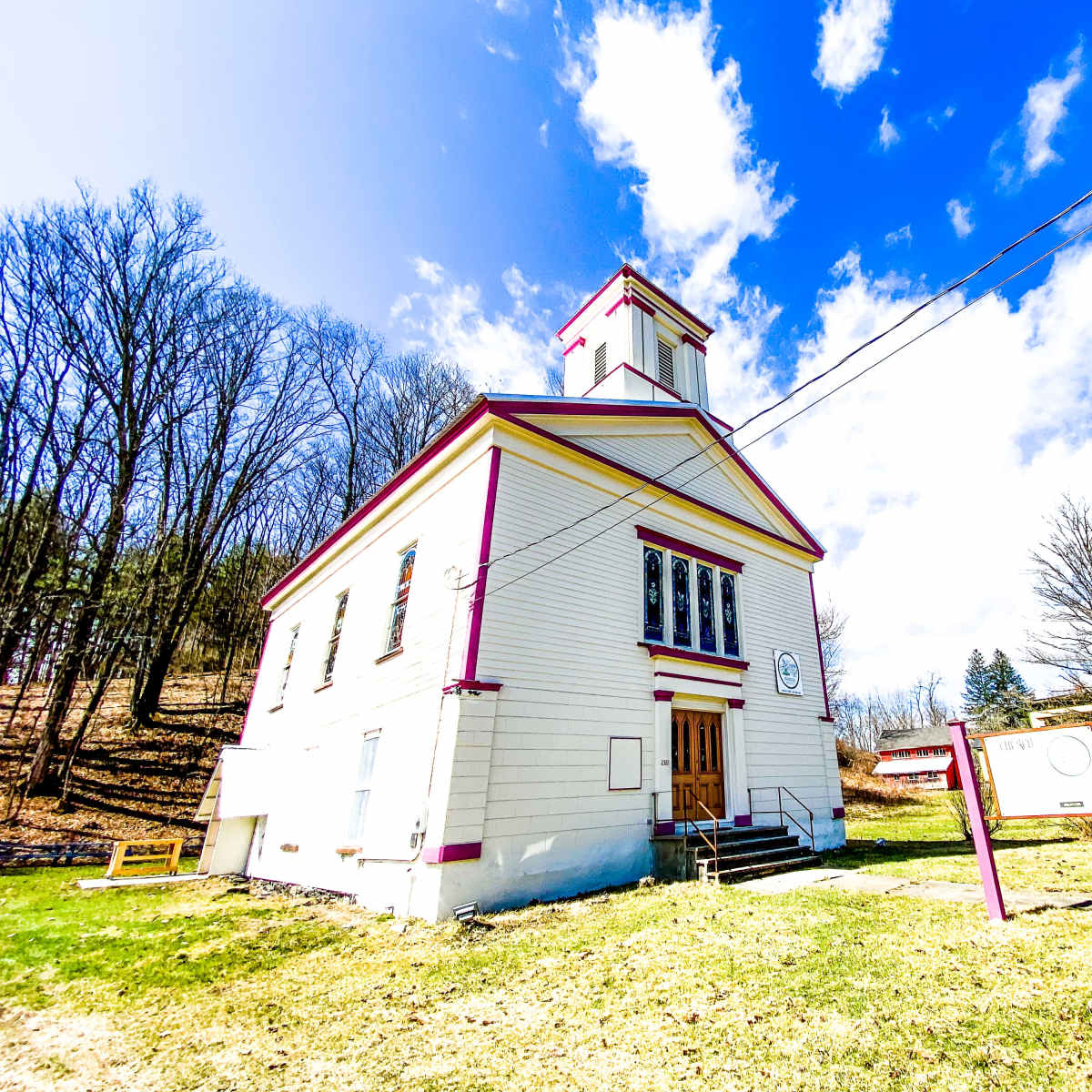 A white, two-story building with purple trim and a bell tower stands beside a road, surrounded by sparse trees and a blue sky with fluffy clouds.