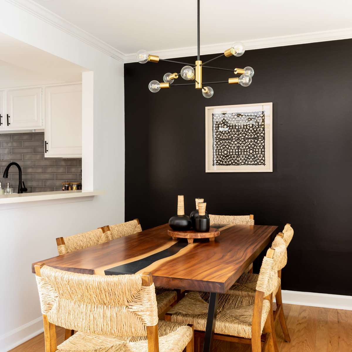 A dining area features a wooden table surrounded by woven chairs, with a black accent wall adorned by a framed artwork and a modern light fixture overhead.