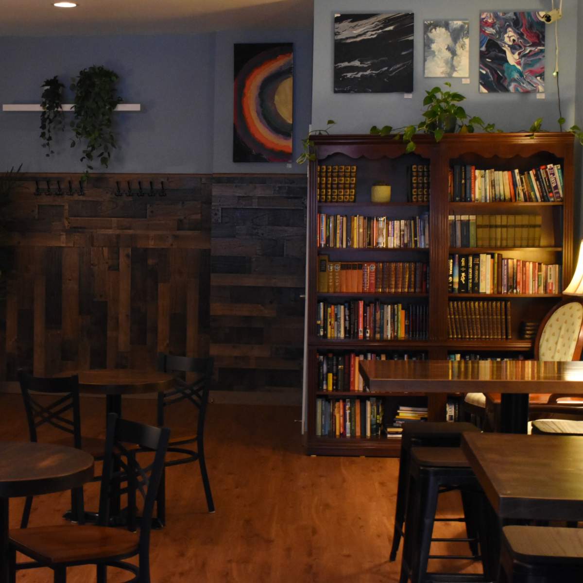 A cozy café interior with wooden tables, a bookshelf filled with books, and colorful artwork displayed on the walls.