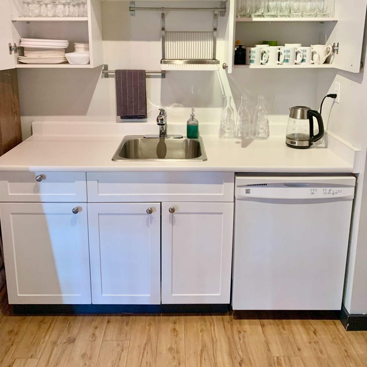 A compact kitchen area with a sink, countertop, dishwasher, and upper shelves filled with glassware and dinnerware.