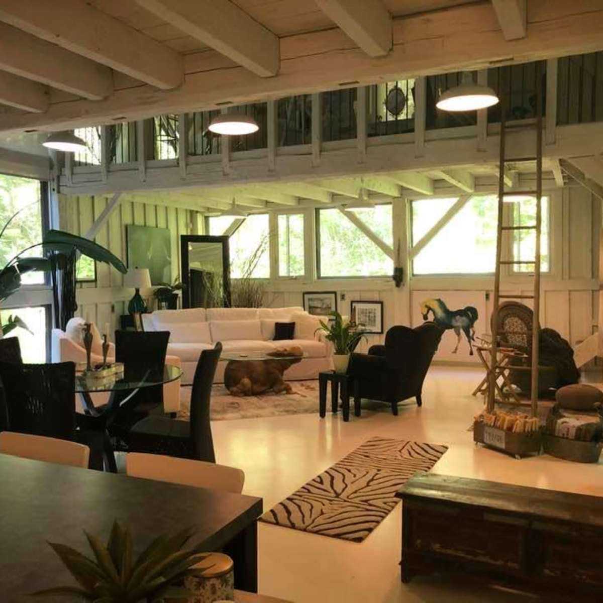 A well-lit, open-concept living space featuring a white sectional sofa, a dining area with a glass table, and large windows overlooking a green landscape.