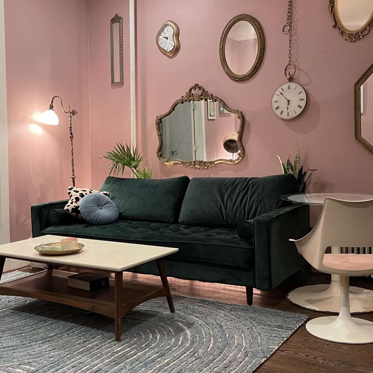 A stylish living room with a green sofa, a round coffee table, two white chairs, and decorative mirrors on pink walls.