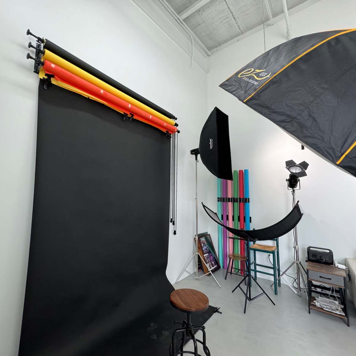 A photography studio with a black backdrop, colorful backdrop rolls, various lighting equipment, and a seating area.