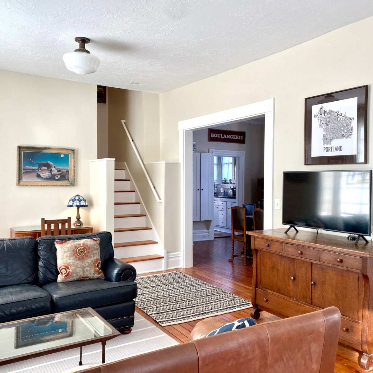 A living room features a dark leather sofa, a glass-top coffee table, a wooden sideboard with a television, and a staircase leading to the upper level.
