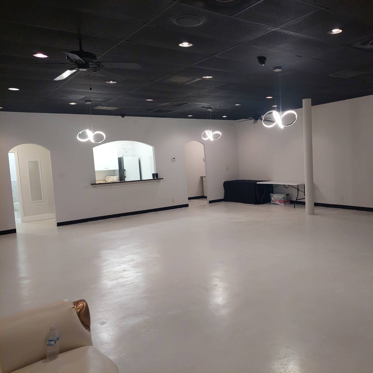 A spacious, empty room with a white floor, black ceiling, and modern light fixtures, along with a small bar area in the background.