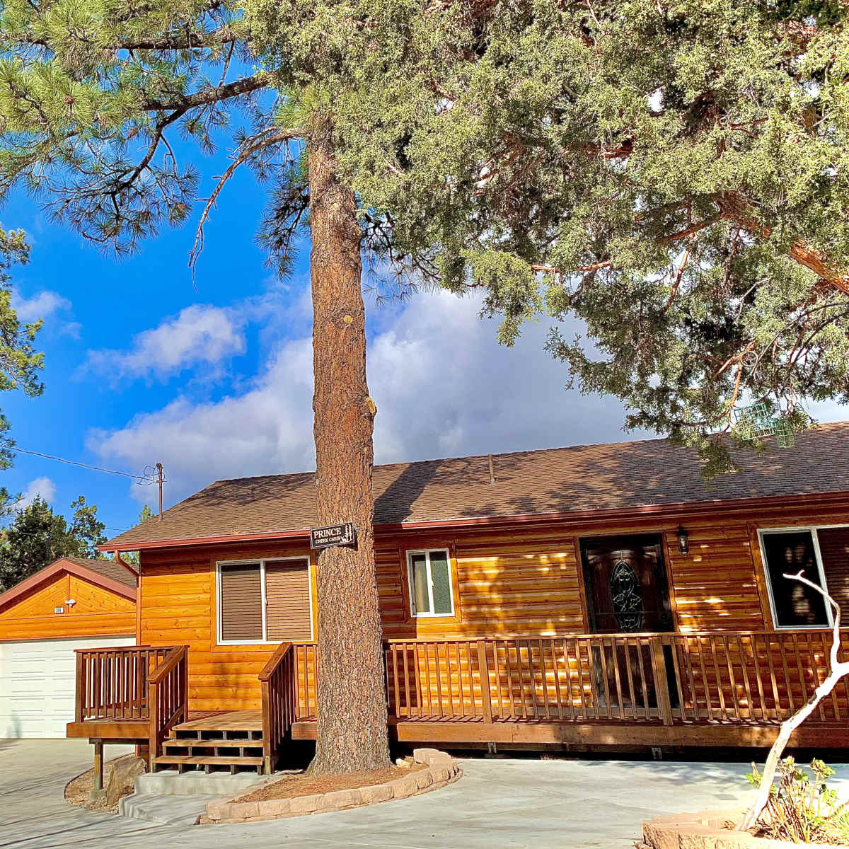 A brown wooden cabin with a porch and a garage is surrounded by tall green trees under a partly cloudy sky.