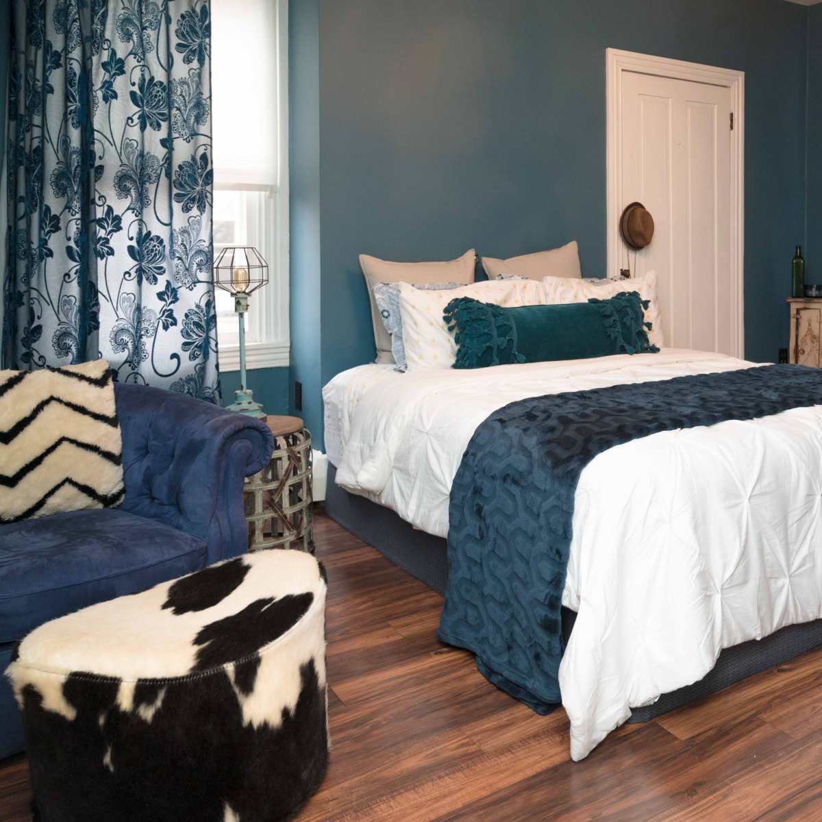 A cozy bedroom with a bed covered in white and blue bedding, a dark blue accent wall, and various decorative furnishings including a patterned chair and a cowhide ottoman.