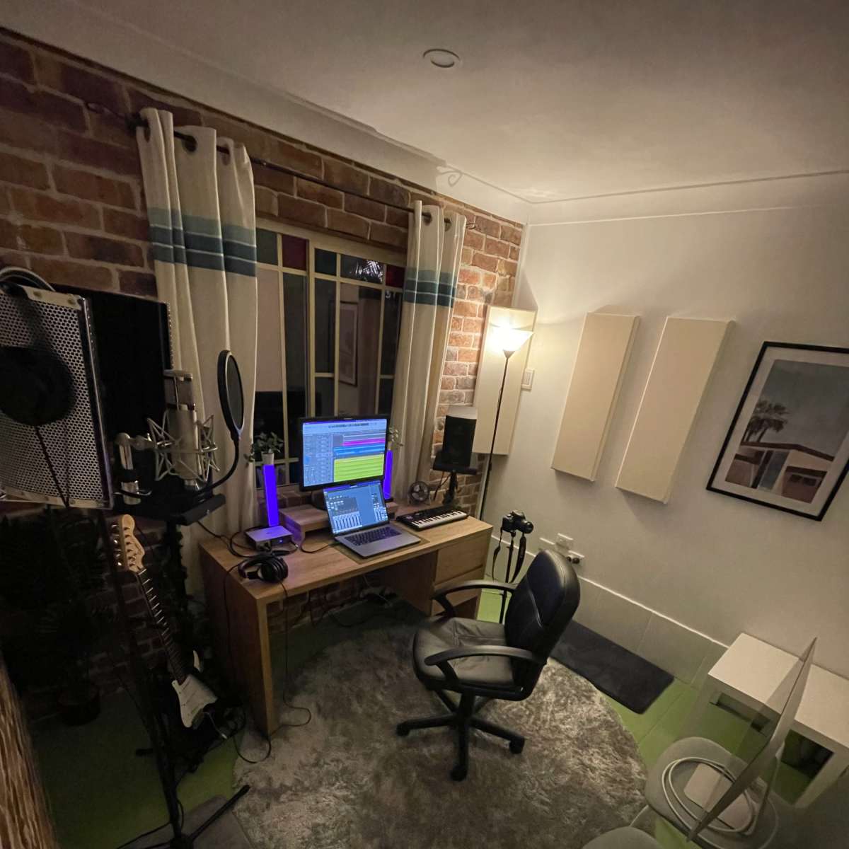A small music production studio featuring a desk with a computer, a microphone setup, and various instruments arranged around a rug.