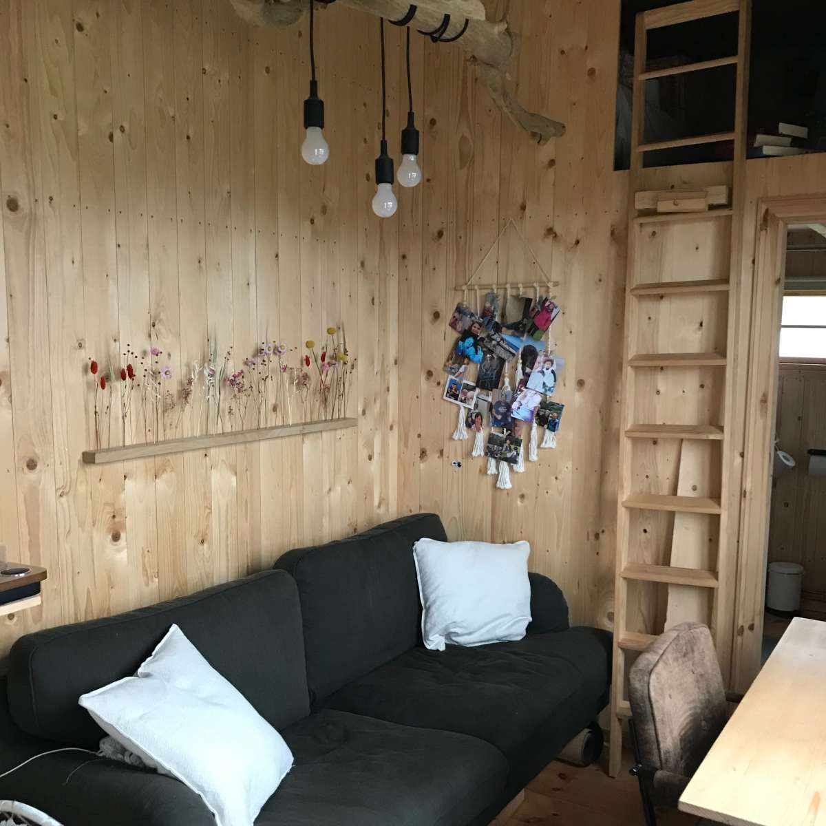 A cozy room with a wooden interior featuring a dark sofa, a small wooden shelf with flowers, a ladder leading to an upper level, and a desk with a chair.