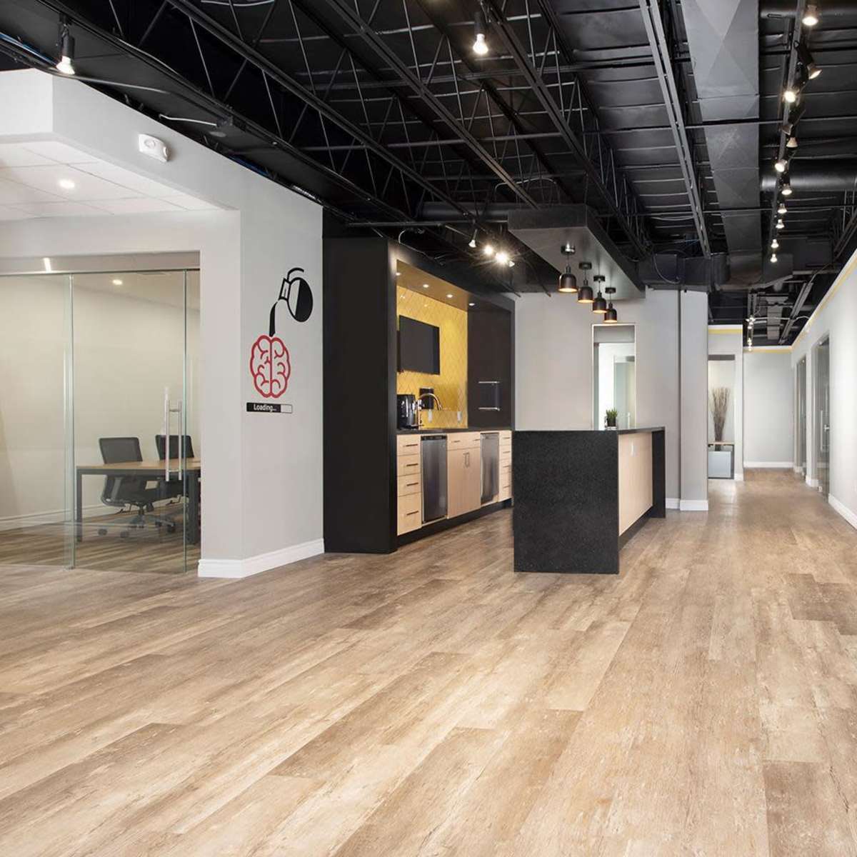 A modern office interior with a kitchen area on one side and glass-walled meeting rooms along the hallway.