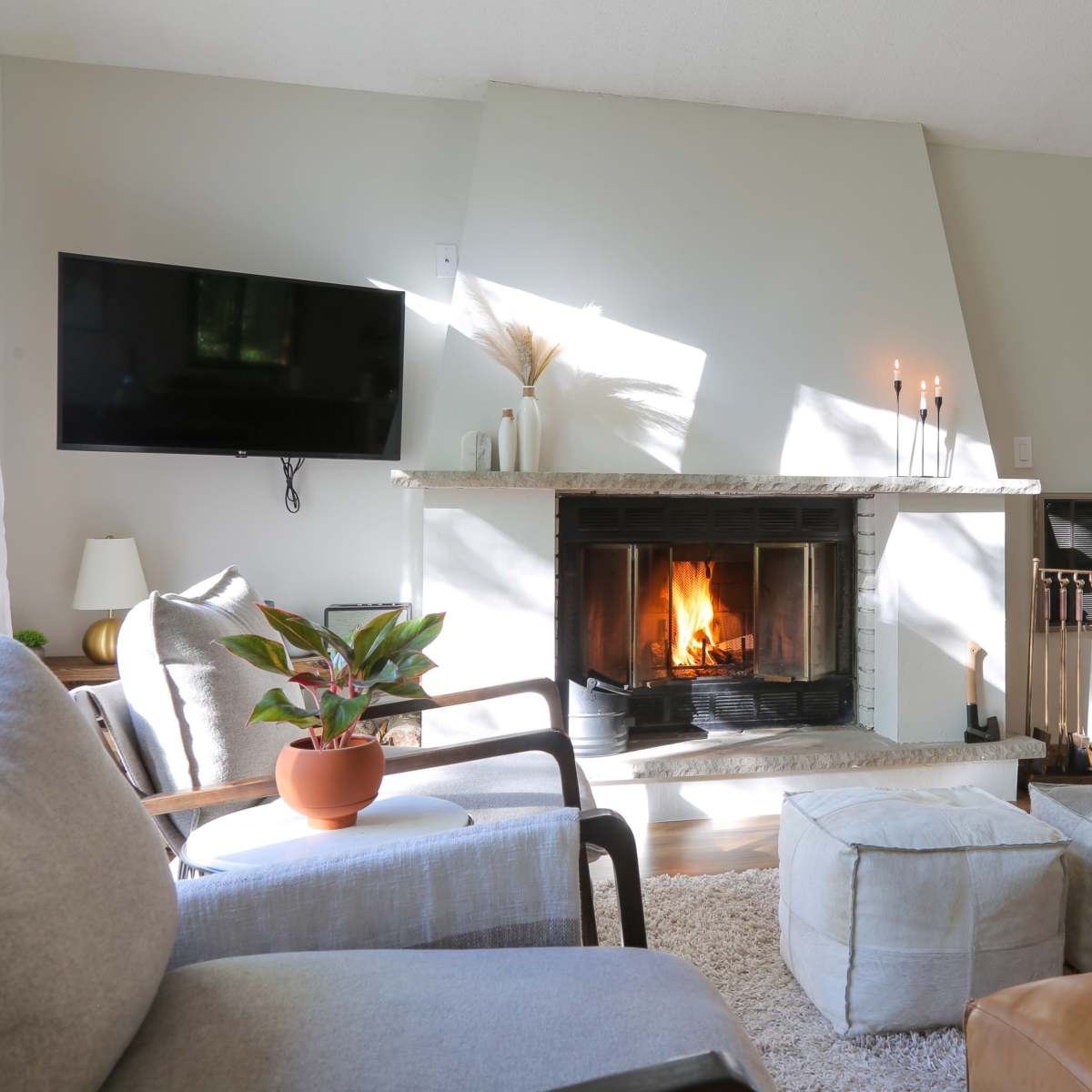 A cozy living room features a stone fireplace with a lit fire, a television mounted on the wall, and comfortable seating arrangements, including a gray armchair and a brown leather couch.