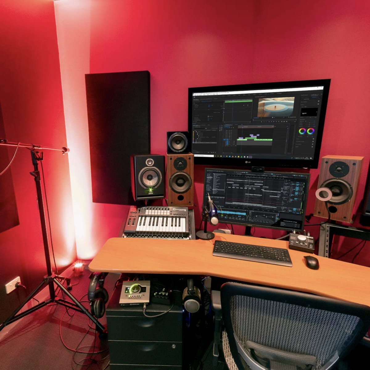 A music production studio featuring a microphone, computer monitor, speakers, and a keyboard arranged on a wooden desk against red-lit walls.
