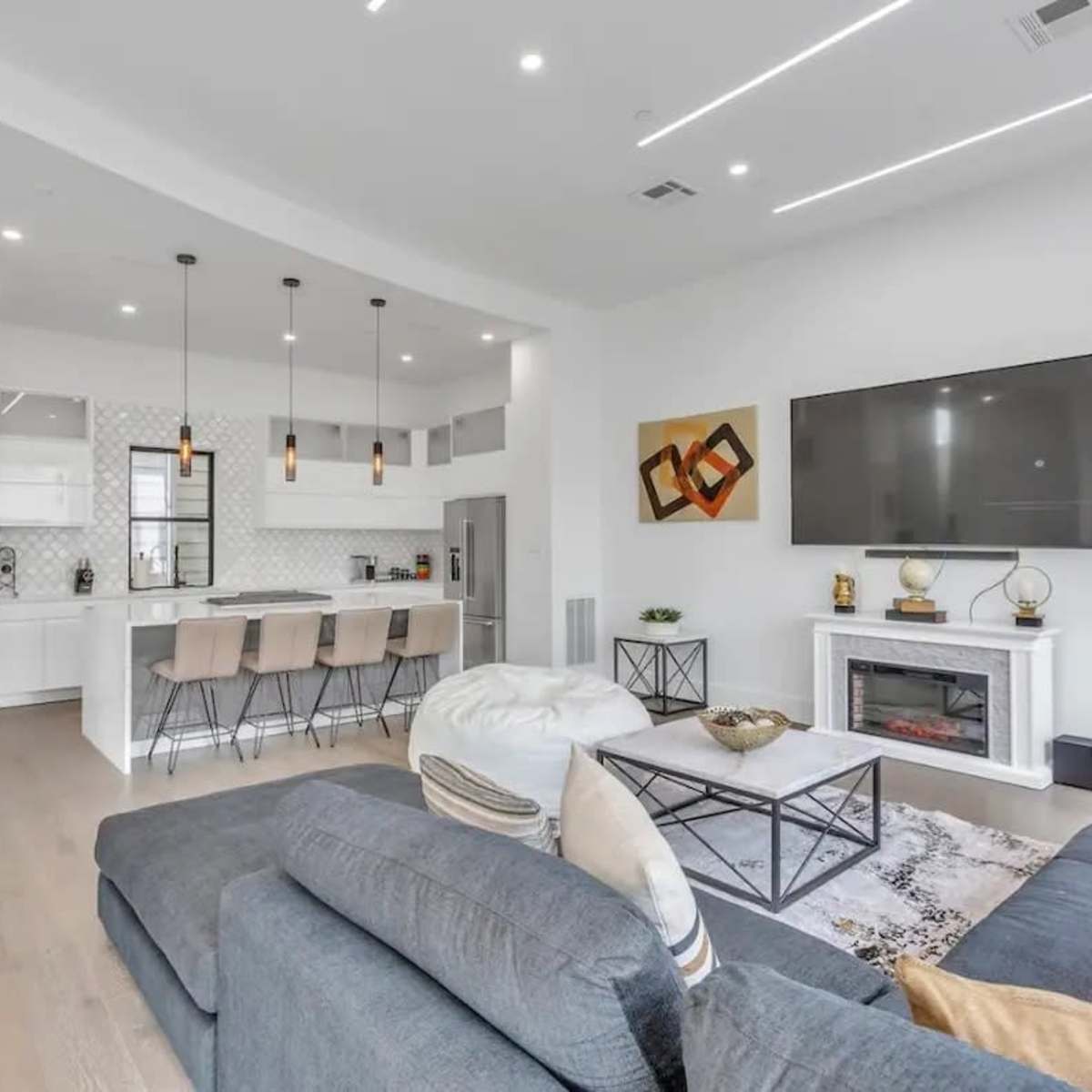 A modern living room featuring a large gray sectional sofa, a flat-screen television mounted on the wall, and a kitchen area with bar stools and pendant lighting in the background.