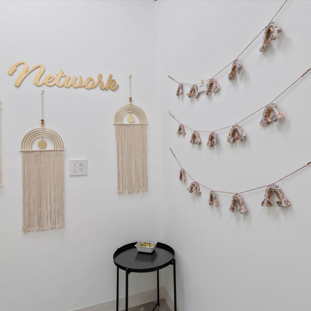 A corner of a room with woven wall hangings and decorative garlands, alongside a small black table.