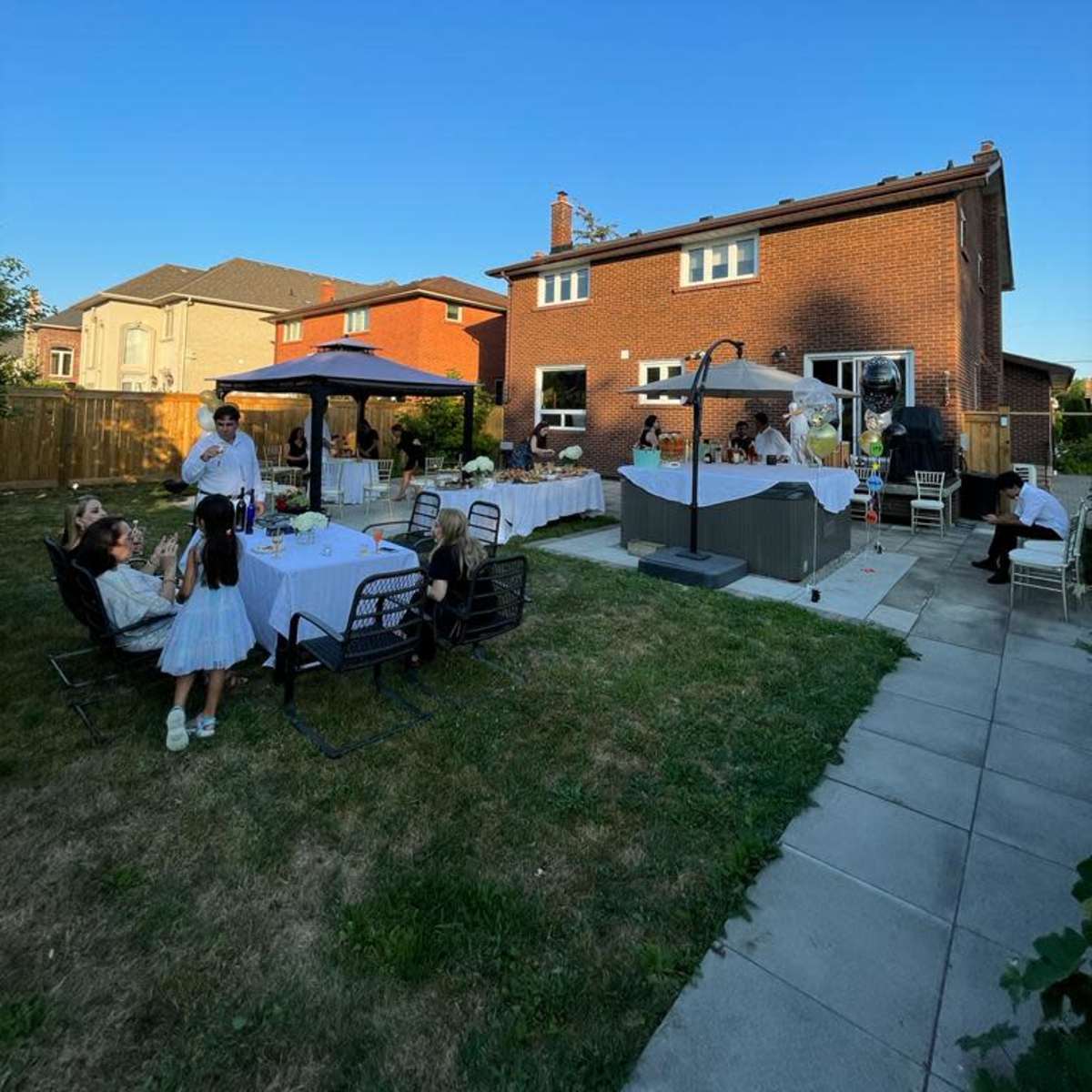 A backyard gathering features tables set for dining, a gazebo, and guests socializing near a house with a clear blue sky above.