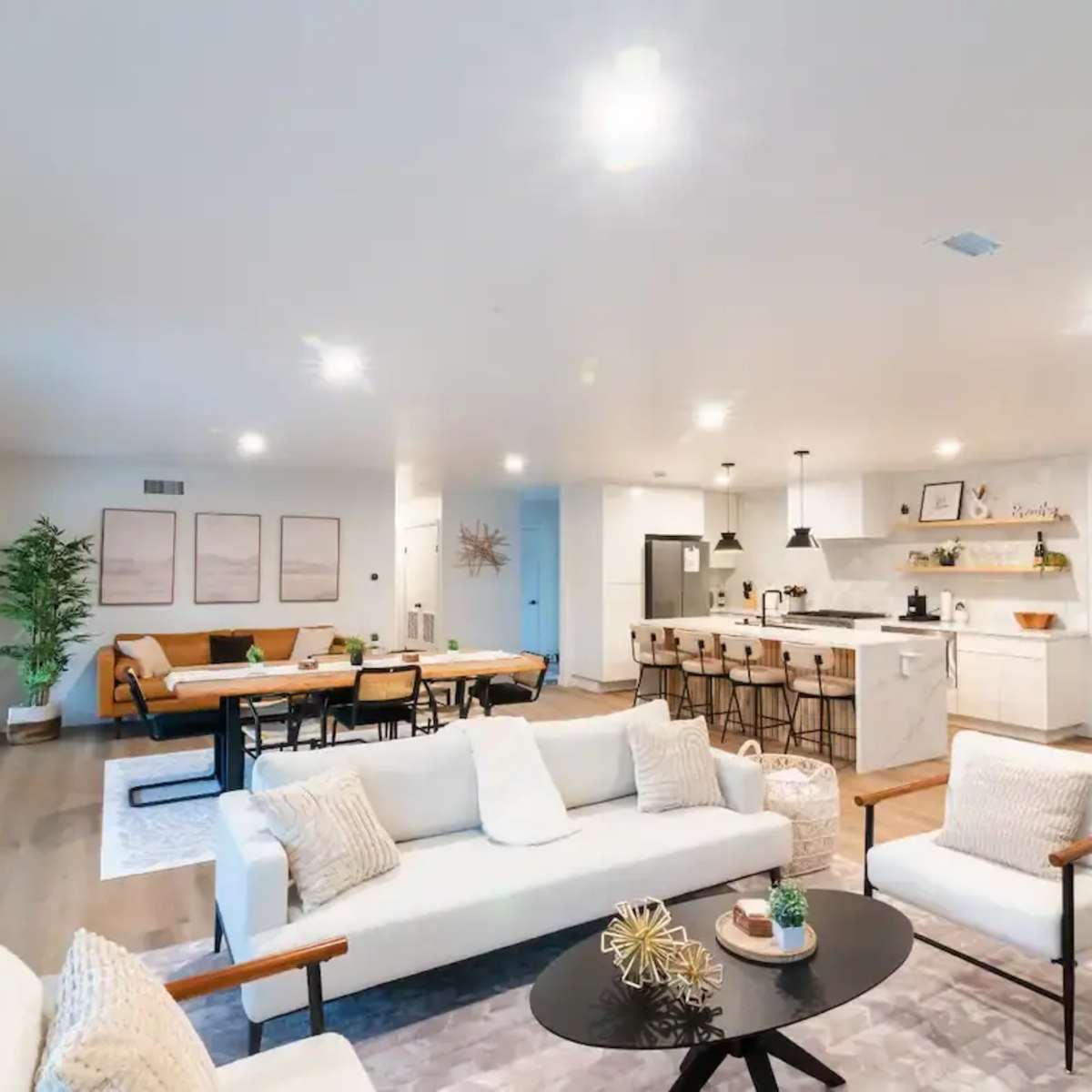A modern open-concept living room and kitchen with a light-colored sofa, a central coffee table, a dining table, and bar seating.