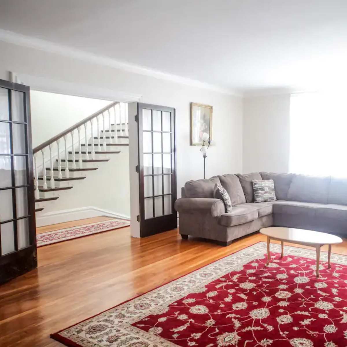 A spacious living room features a sectional sofa, a coffee table, and a large window illuminating the space, with a staircase and entryway visible in the background.