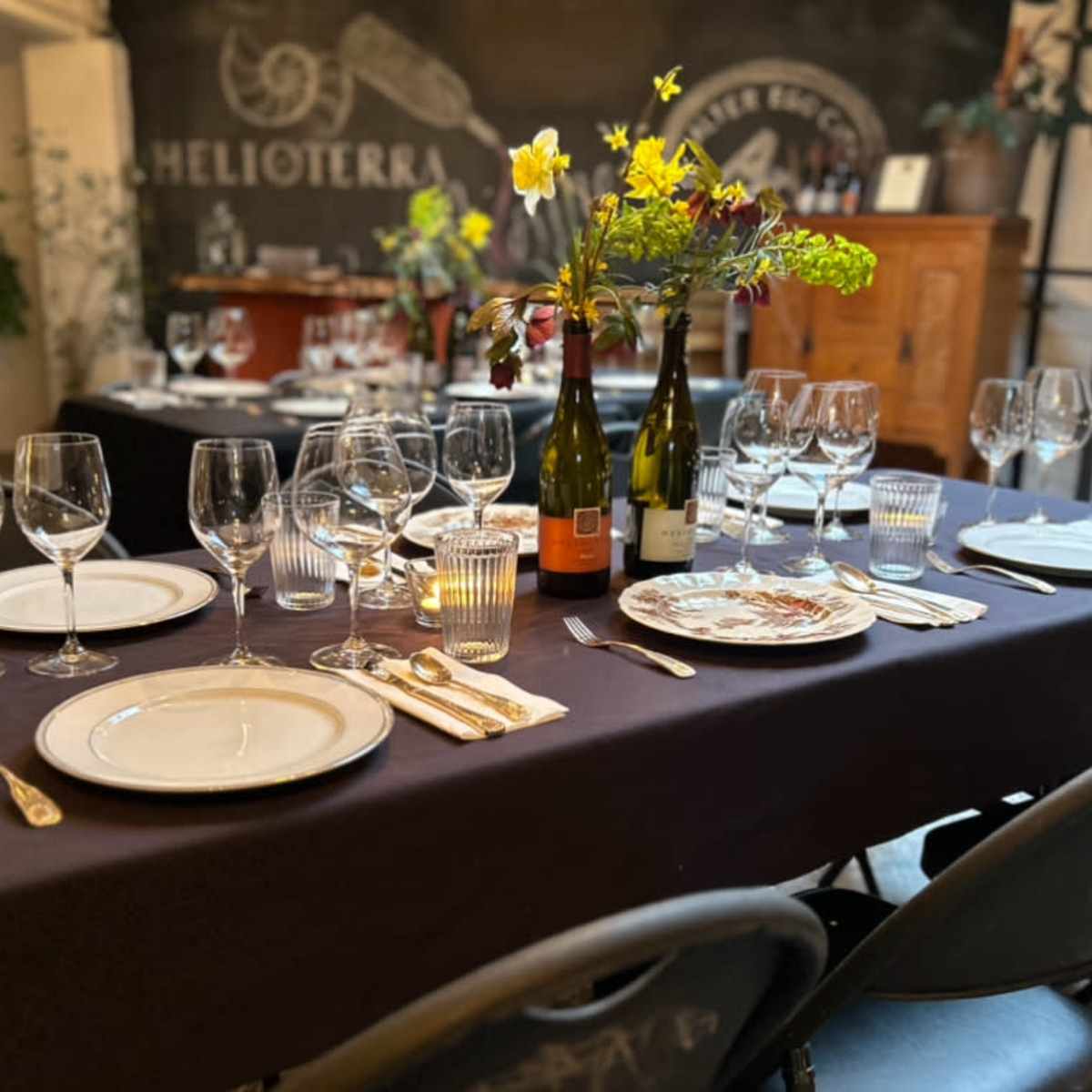 A dining table is set with plates, wine glasses, and utensils, surrounded by bottles of wine and decorative flowers, ready for a gathering.