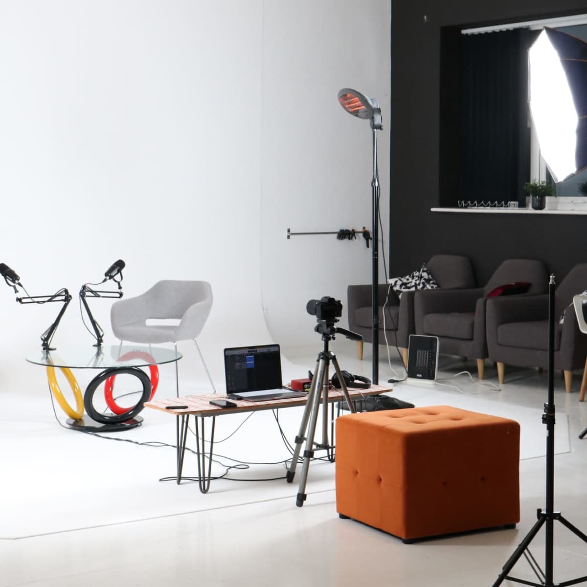 A modern studio setup features a glass table, a gray chair, a tripod with a camera, and various lighting equipment.