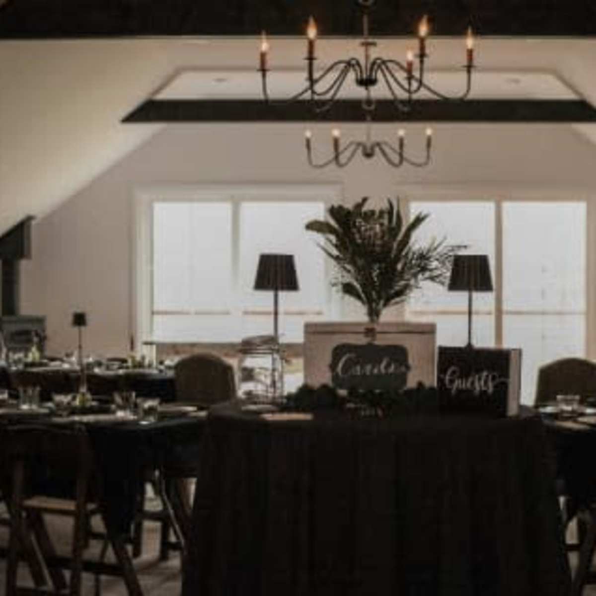 A dining area features neatly arranged tables with dark tablecloths, chairs, and a centerpiece under a chandelier.