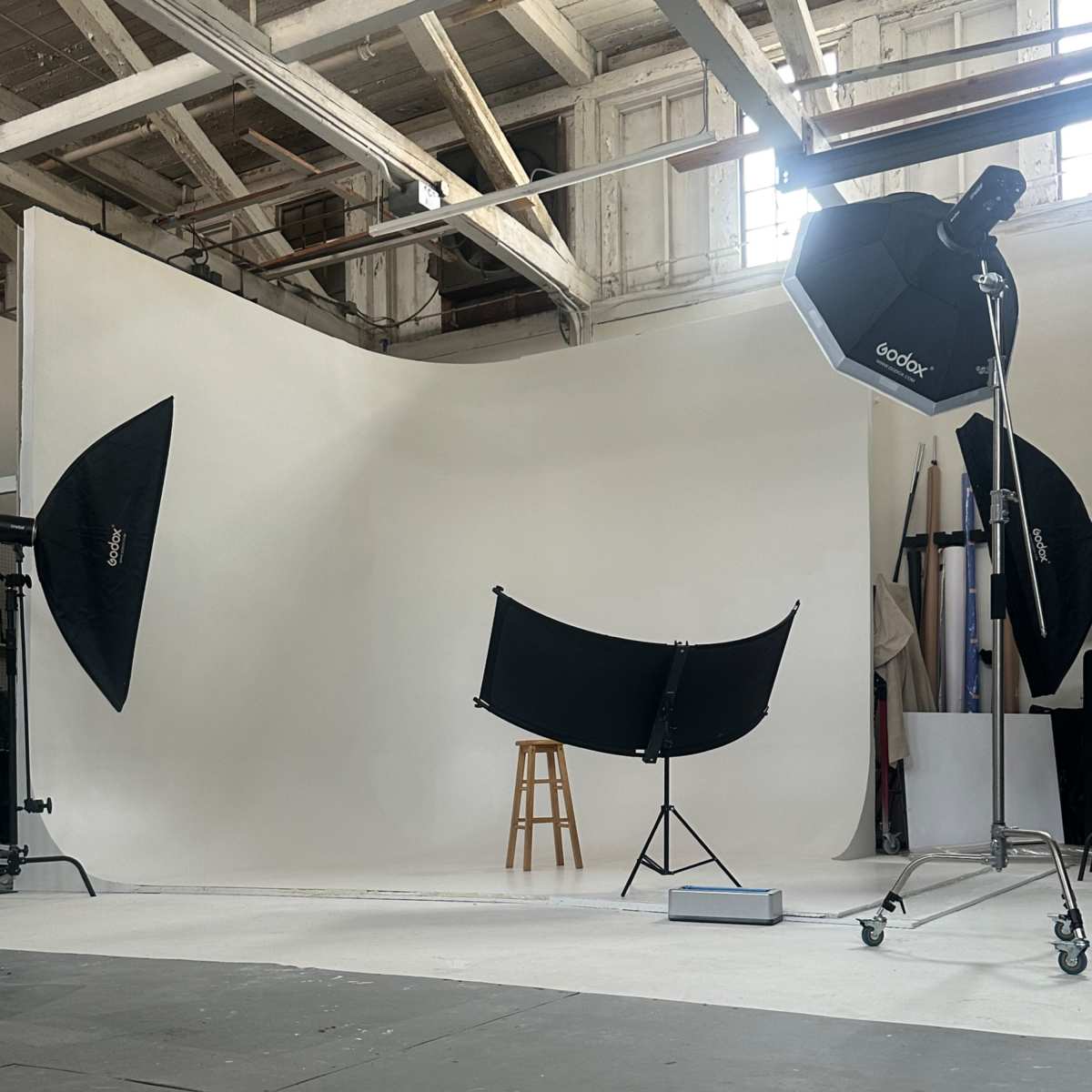 A photography studio with a plain backdrop, a wooden stool, and studio lighting equipment positioned around the setup.
