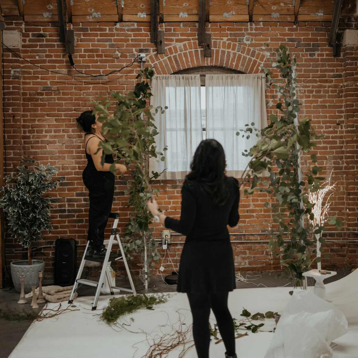 Two people are arranging greenery and decorative elements in a rustic, brick-walled space for an event.