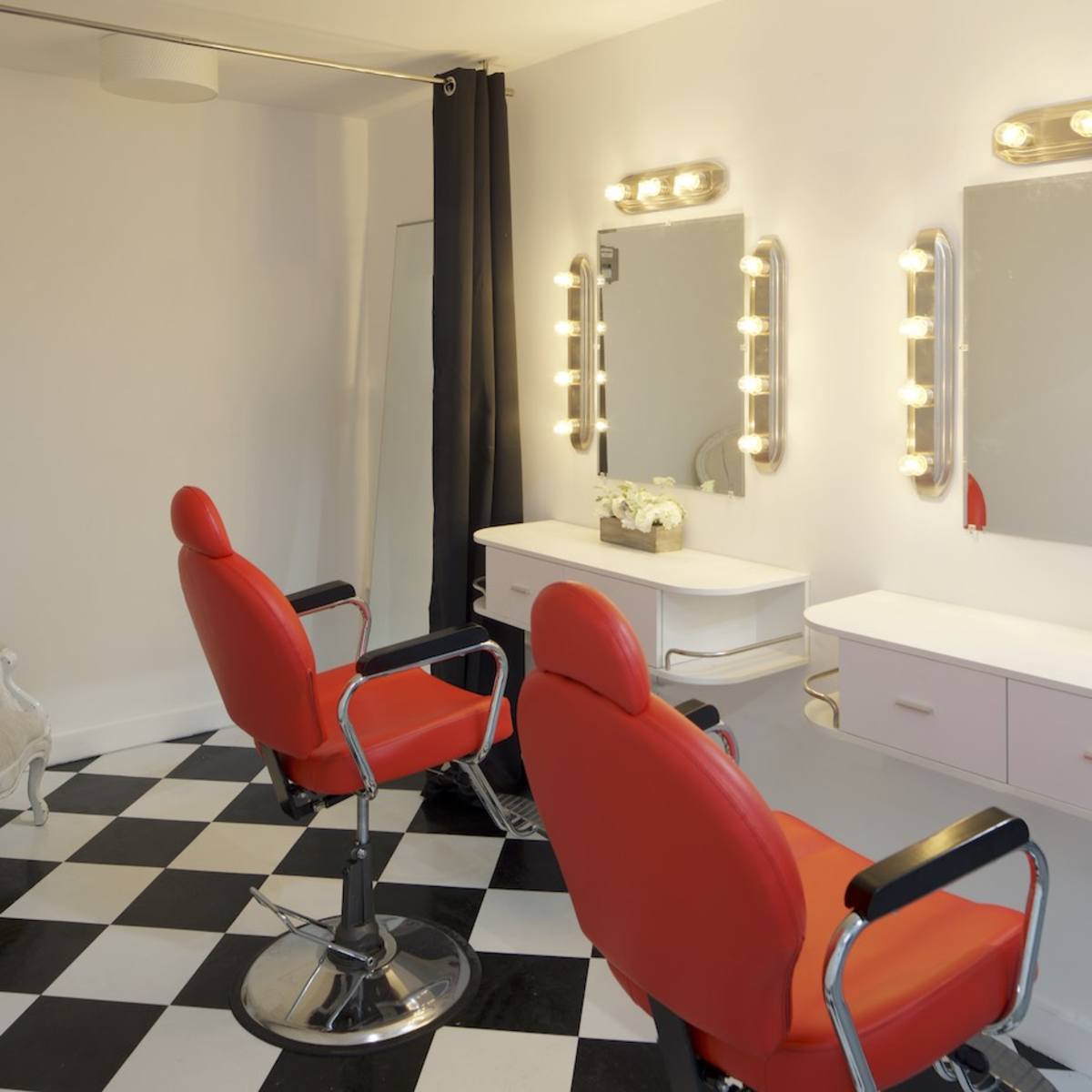 A hair salon with two red barber chairs, a checkered floor, and illuminated mirrors against a white wall.