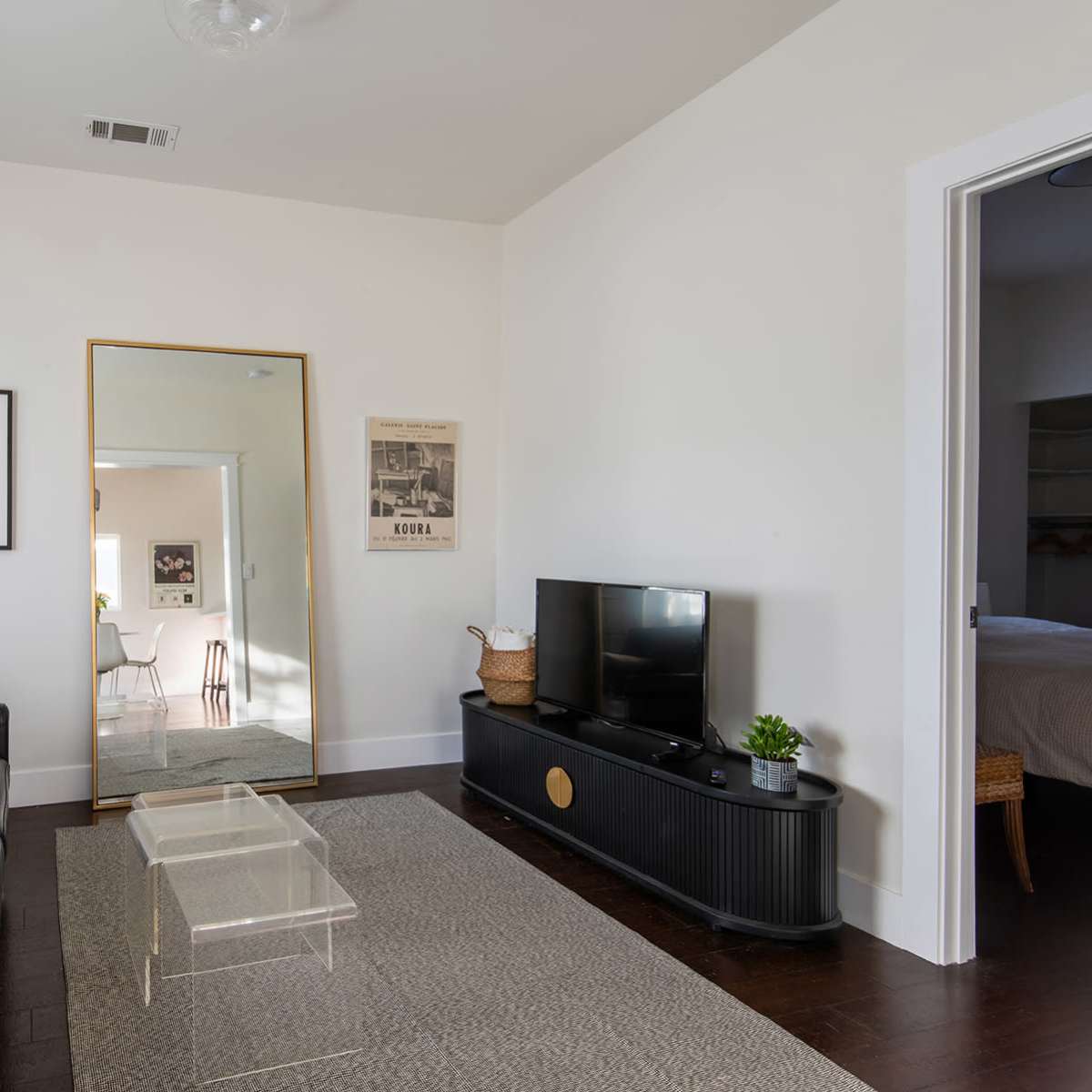 A living room features a black sofa, a glass coffee table, a large mirror, and a television on a stand, with light streaming through the windows.