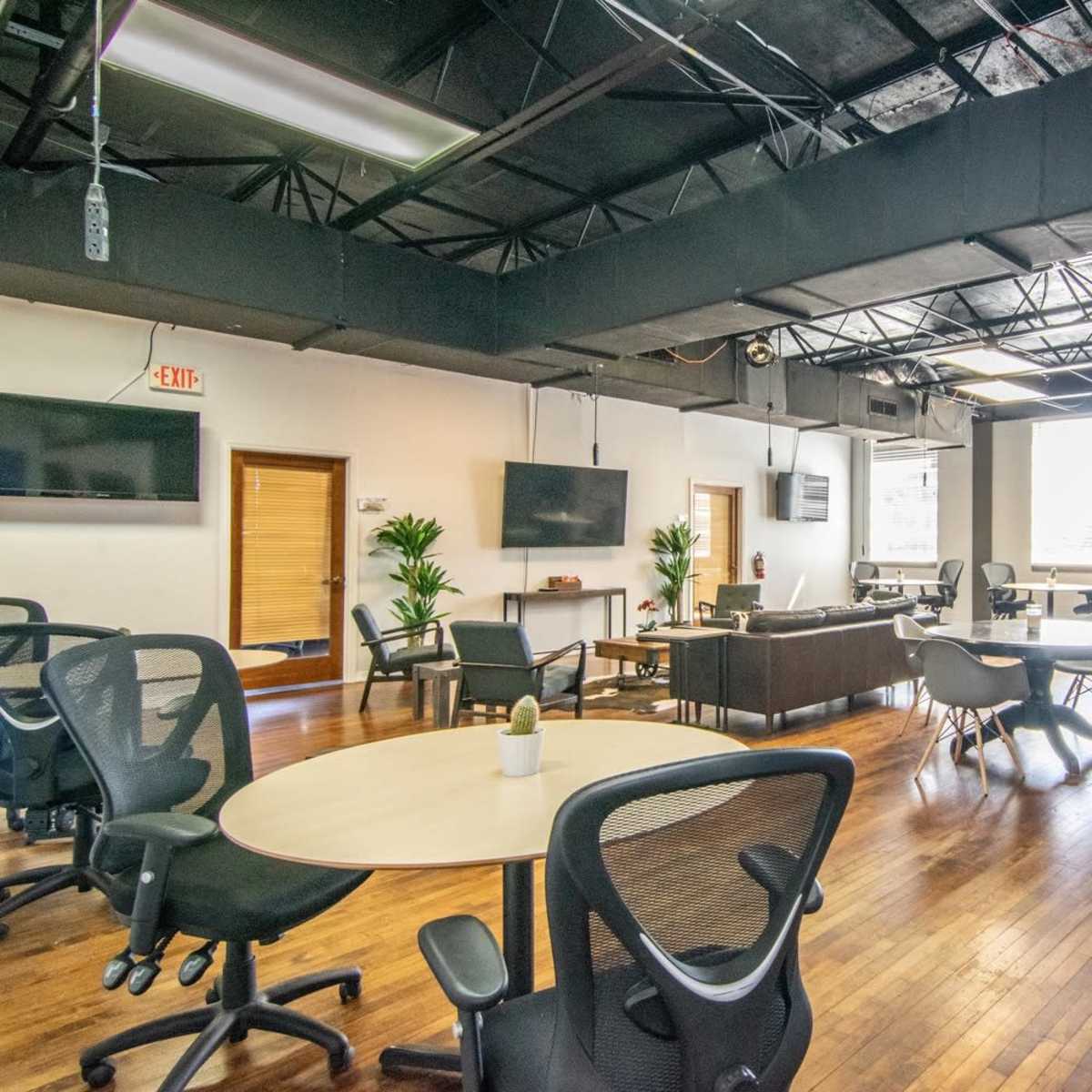 An open workspace with several tables and chairs, large windows, and a couch area, featuring wooden flooring and modern furnishings.