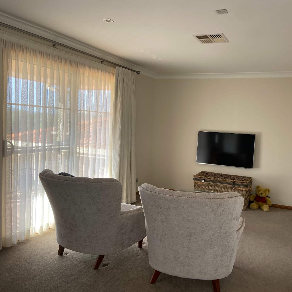 The room features two upholstered armchairs facing a wall-mounted television, with large curtains covering a window and a teddy bear sitting on a nearby storage basket.