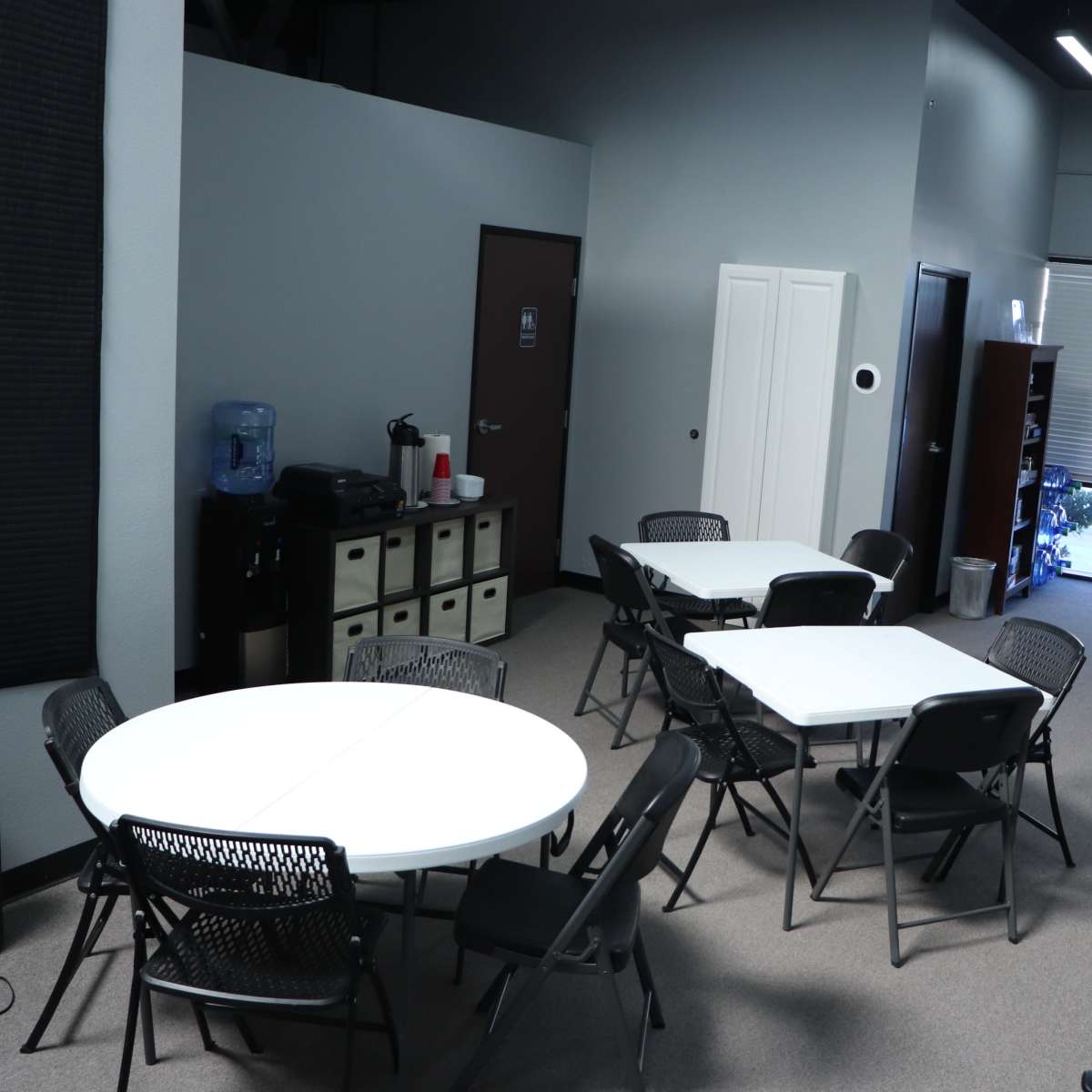 An interior space with several round tables and chairs arranged on a carpeted floor, alongside a water cooler and storage cabinets.